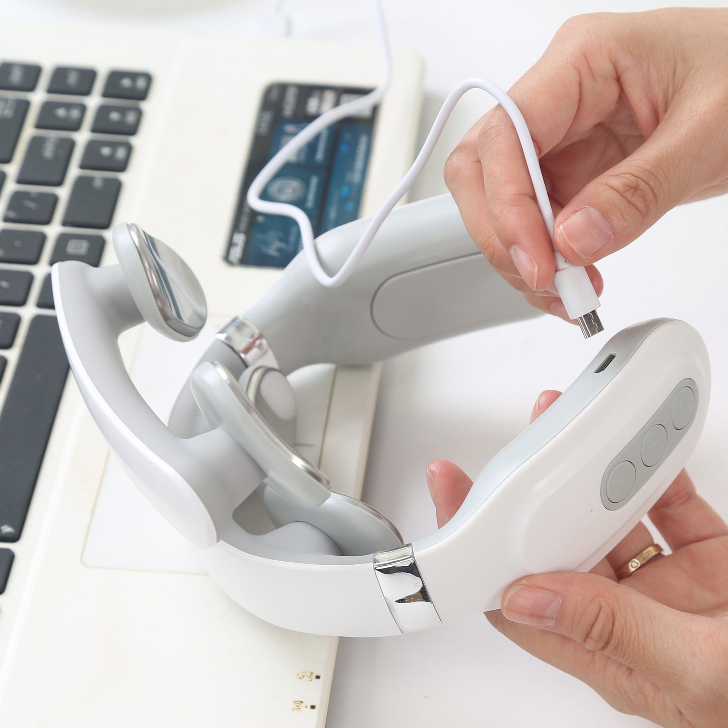 Hand holding a white and gray electronic neck massager with a laptop and card in the background